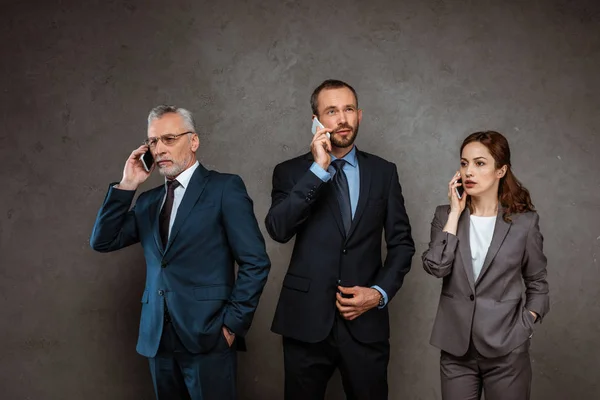 Handsome businessmen and attractive businesswoman in formal wear talking on smartphones on grey — Stock Photo