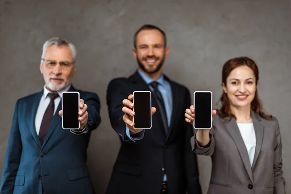 Избирательный фокус жизнерадостных бизнесменов и бизнесвумен, держащих смартфоны с пустыми экранами на сером — стоковое фото