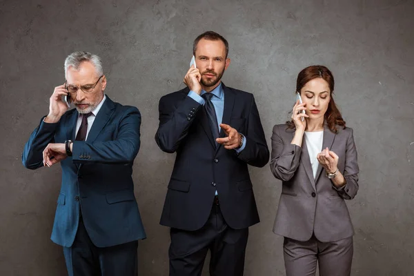 Hombres de negocios guapos hablando en el teléfono inteligente y señalando con el dedo mientras mira a la cámara cerca de los socios en gris - foto de stock