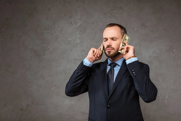 Hombre guapo sosteniendo billetes de dólar cerca de las orejas mientras está de pie con los ojos cerrados en gris - foto de stock