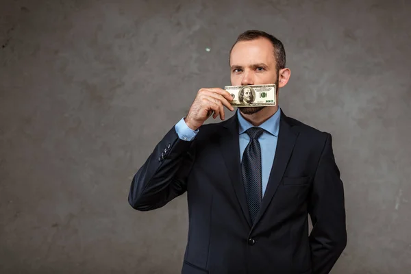 Бизнесмен в костюме, прикрывающий рот банкнотой в долларах, стоя на сером — стоковое фото