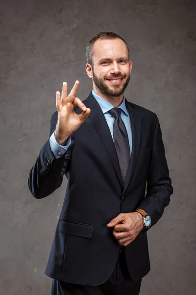 Happy bearded businessman in suit showing ok sign on grey — Stock Photo