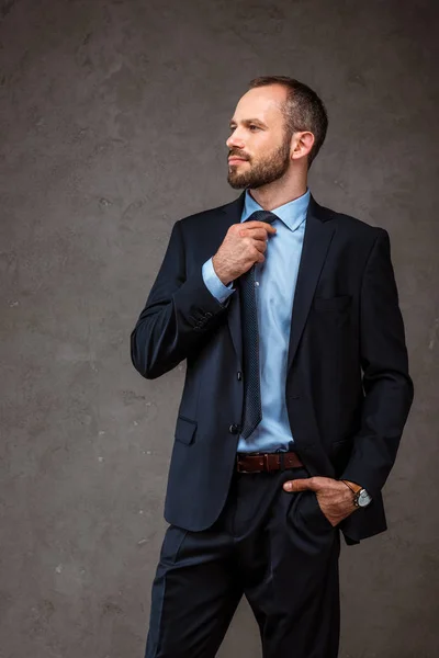 Bonito empresário de pé com mão no bolso e tocar gravata no cinza — Fotografia de Stock