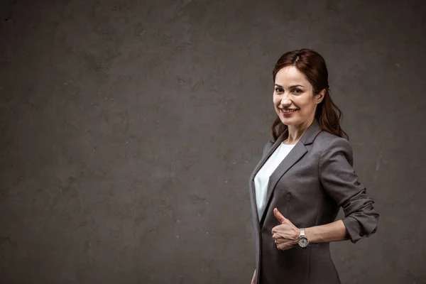 Cheerful brunette businesswoman showing thumb up and smiling on grey — Stock Photo