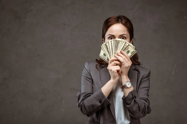 Shocked brunette businesswoman covering face with dollar banknotes on grey — Stock Photo