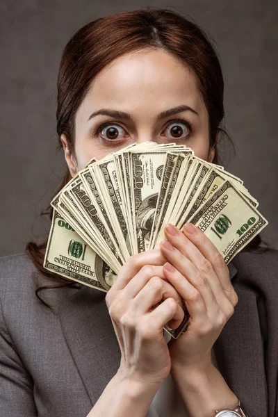 Femme brune choquée couvrant le visage avec des billets en dollars sur gris — Photo de stock