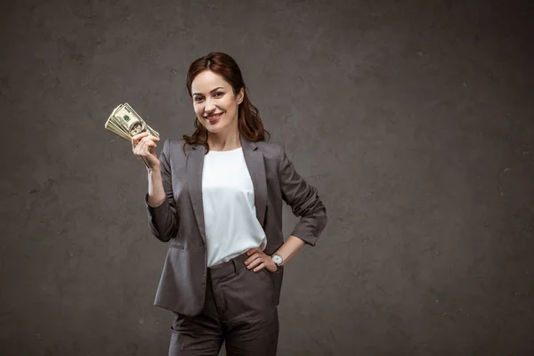 Happy businesswoman standing with hand on hip and holding dollar banknotes on grey — Stock Photo