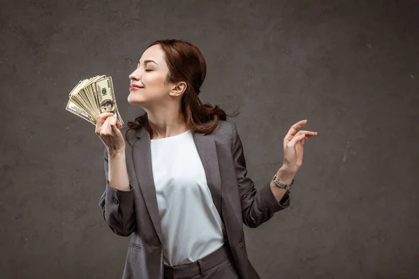 Веселая деловая женщина с закрытыми глазами, пахнущая долларовыми банкнотами на сером — стоковое фото