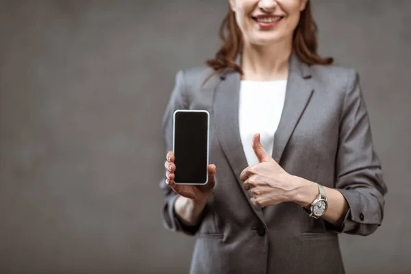 Selektiver Fokus der fröhlichen Geschäftsfrau, die den Daumen nach oben zeigt und ihr Smartphone mit leerem Bildschirm auf grau hält — Stockfoto