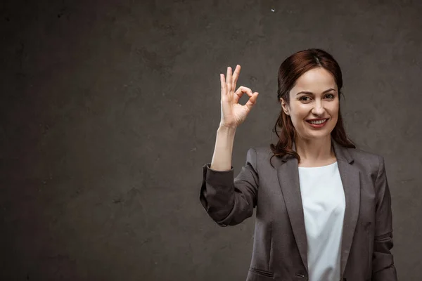 Cheerful brunette woman showing ok sign and smiling on grey — Stock Photo