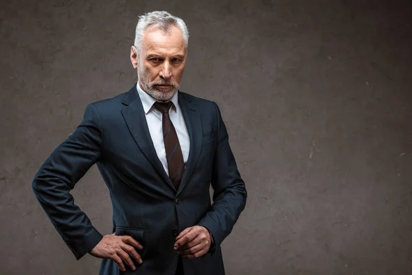 Hombre de negocios confiado en traje de pie con la mano en la cadera en gris - foto de stock