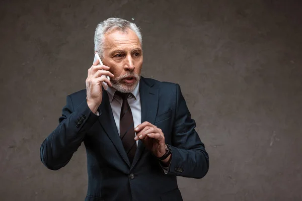 Surprised bearded businessman talking on smartphone on grey — Stock Photo