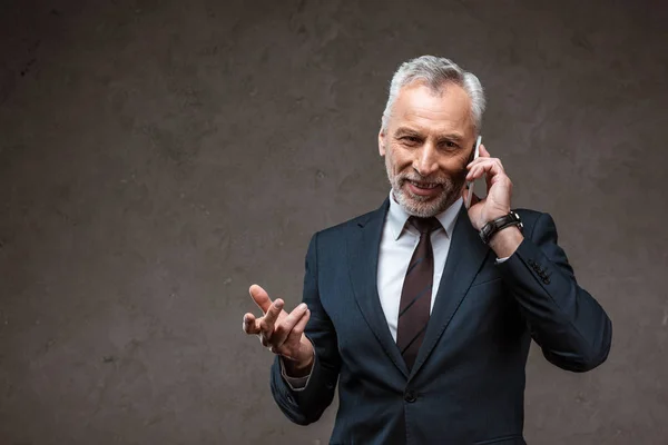 Alegre hombre de negocios en traje hablando en el teléfono inteligente y el gesto en gris - foto de stock