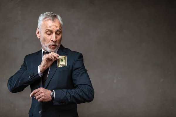 Bearded businessman in suit putting dollar banknote in pocket on grey — Stock Photo