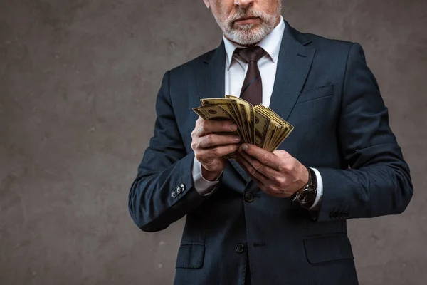 Vue recadrée de l'homme d'affaires en costume tenant des billets en dollars dans les mains sur gris — Photo de stock