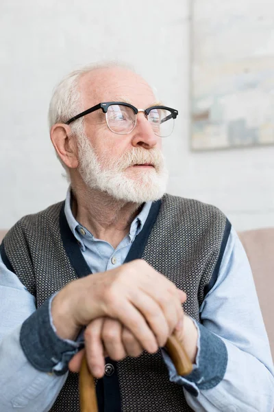 Uomo anziano calmo e triste con bastone da passeggio che distoglie lo sguardo, seduto in una stanza luminosa — Foto stock