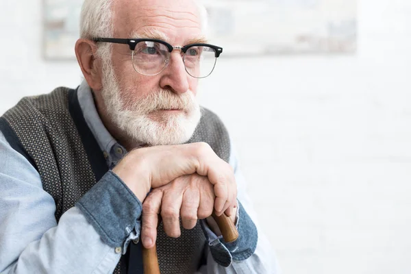 Sad and bearded senior man with walking stick sitting in room, looking away — Stock Photo