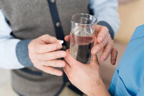 Обрізаний вид пацієнта тримає таблетки в руці, а жінка дає склянку з водою літньому чоловікові — стокове фото