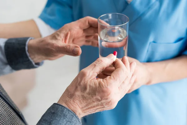 Vue recadrée de l'homme âgé tenant des pilules dans la main, tandis que le médecin donnant du verre avec de l'eau au patient — Photo de stock