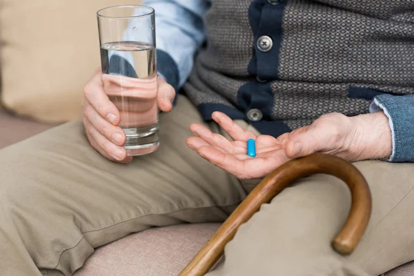 Cropped view of elderly man holding pill and glass with water in hands, sitting on couch — Stock Photo