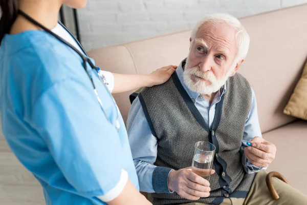 Nurse putting hands on grey haired man with pill and glass of water — Stock Photo