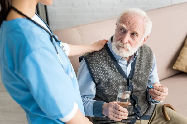 Krankenschwester legt grauhaarigen Mann mit Tablette und Glas Wasser die Hand auf — Stockfoto