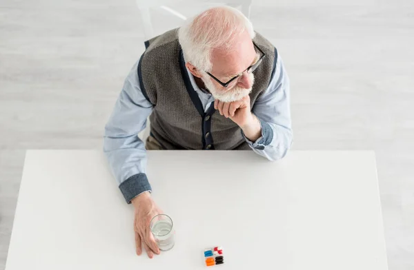Вид сверху на задумчивого человека, смотрящего в сторону, сидящего за столом с таблетками и водой в стекле — стоковое фото