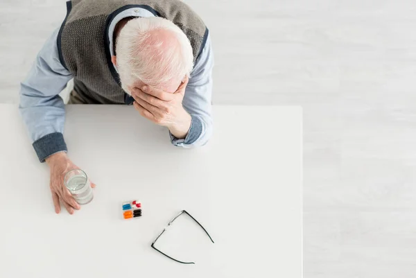 Overhead view of upset elderly man sitting behind table with pills and water in glass — Stock Photo