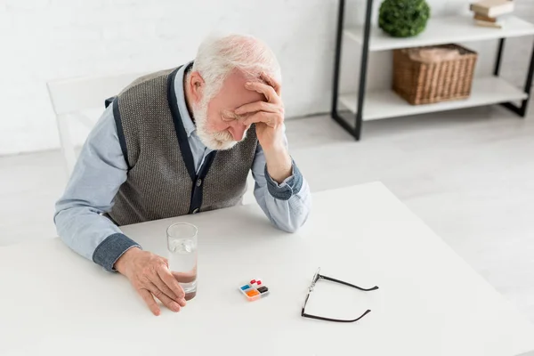 Vista de ángulo alto del hombre mayor molesto sentado detrás de la mesa con pastillas, cogido de la mano en la cabeza - foto de stock