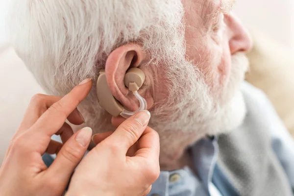 Обрізаний вигляд жінки, що допомагає сірому волосистому чоловікові, в слуховому апараті — стокове фото