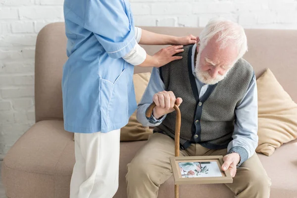 Nurse putting hands on upset and grey haired man with photo frame — Stock Photo