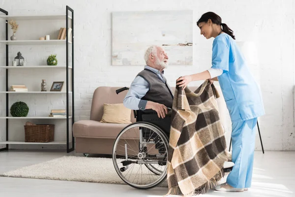 Vista lateral de la enfermera cuidadora, cubriendo manta discapacitada y hombre de pelo gris en silla de ruedas - foto de stock