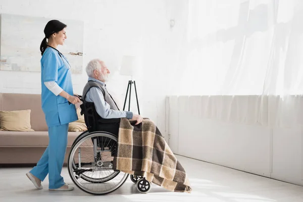 Vue latérale de l'infirmière heureuse debout dans la chambre avec espace de copie, et portant sur handicapé homme aux cheveux gris en fauteuil roulant — Photo de stock
