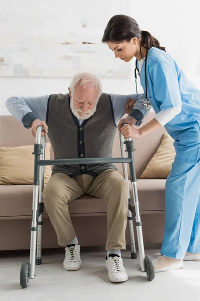 Медсестра помогает пенсионеру, встает с дивана — стоковое фото