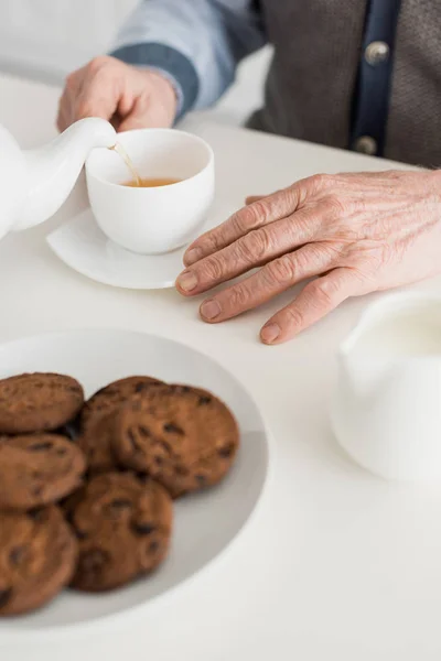 Abgeschnittene Ansicht der Hand eines älteren Mannes auf weißem Tisch mit Teetasse und Keksen — Stockfoto