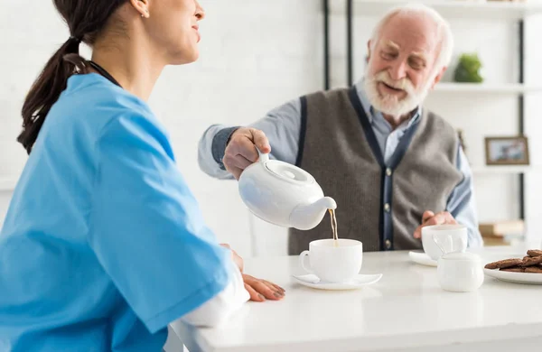 Hombre mayor feliz y sonriente, sirviendo té en taza blanca, sentado en la cocina en casa - foto de stock