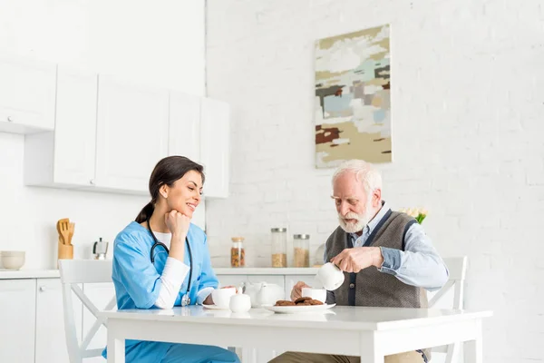 Happy doctor siting on kitchen behind table with senior man — Stock Photo