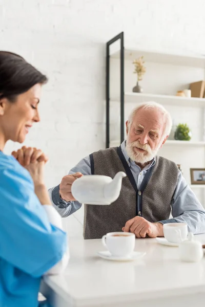 Allegro infermiera seduta sulla cucina a casa, mentre l'uomo anziano versando il tè in tazza — Foto stock