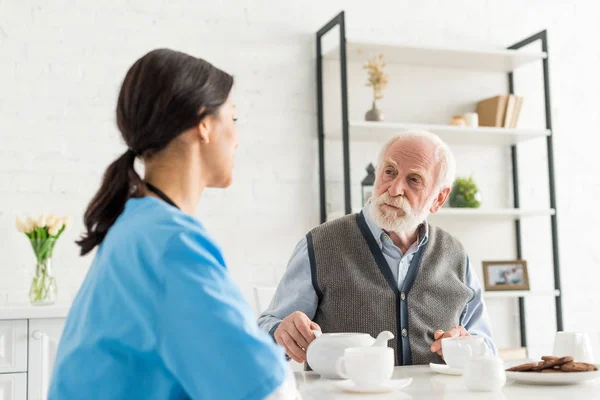 Calm grey haired man looking at nurse, sitting on kitchen at home — Stock Photo