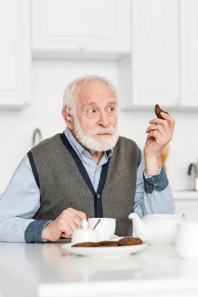 Curioso hombre de pelo gris sentado en la cocina con la galleta en la mano, y mirando hacia otro lado - foto de stock
