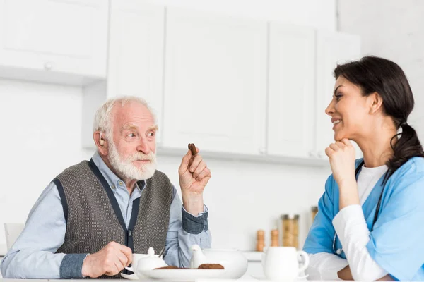 Alegre hombre de pelo gris sentado en la cocina con galletas en la mano, y mirando a la enfermera — Stock Photo