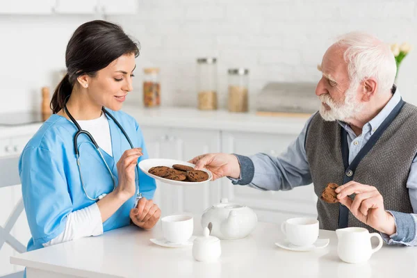 Homem de cabelos grisalhos oferecendo biscoitos para feliz, e enfermeira sorrindo — Fotografia de Stock