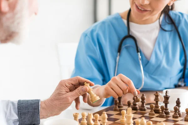 Vista recortada de la enfermera y el hombre mayor sentado en la cocina, y jugando al ajedrez - foto de stock