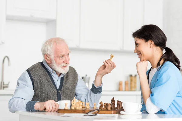 Enfermera y hombre de pelo gris sentado en la cocina brillante, y jugando al ajedrez - foto de stock