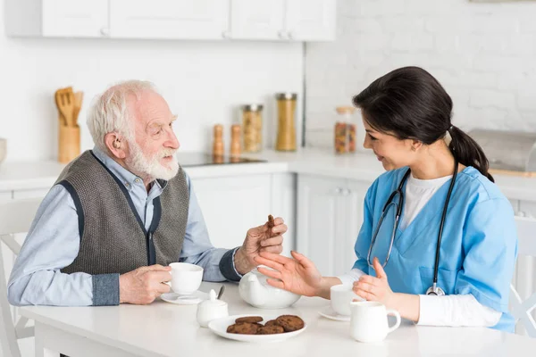 Homme aux cheveux gris avec biscuits et thé assis sur la cuisine, parlant à l'infirmière — Photo de stock