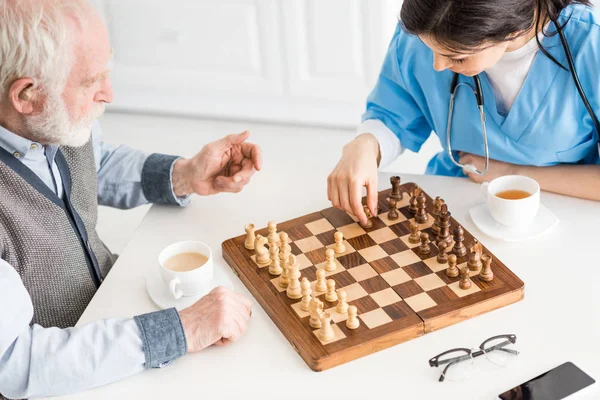 Vista de alto ángulo de la enfermera con el hombre mayor sentado en la cocina, jugando en el ajedrez - foto de stock