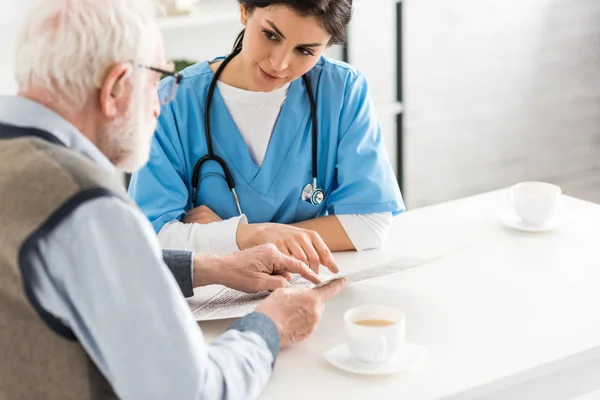 Cropped view of nurse looking at senior man with newspaper in hands — Stock Photo