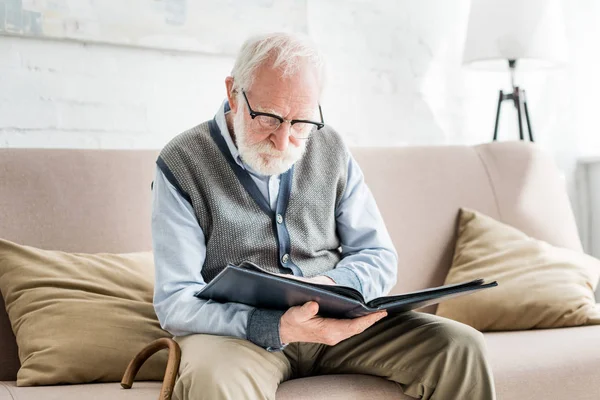 Hombre mayor sentado en el sofá, y sosteniendo álbum de fotos en las manos — Stock Photo