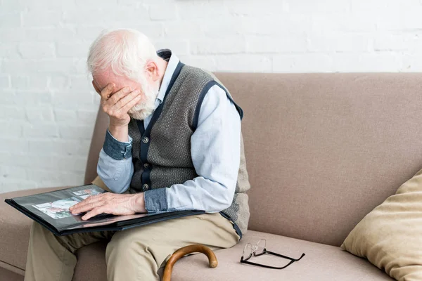 Älterer Mann sitzt auf Sofa, hält Fotoalbum in der Hand und bedeckt sein Gesicht mit der Hand — Stockfoto