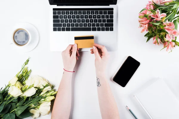 Cropped view of woman holding credit card near laptop, smartphone with blank screen, cup of coffee and bouquets on white — Stock Photo
